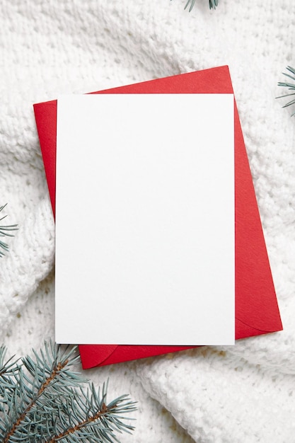 Christmas greeting card mockup with red envelope and fir tree branches on white knitted clothes background top view flat lay