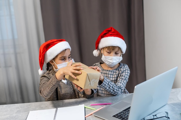 크리스마스 인사말 온라인. 의료용 마스크를 쓴 두 어린 소녀 노트북. 카메라에 선물, 온라인 구매를 보여줍니다. 친구, 친척 및 부모에게 영상 통화 인사말