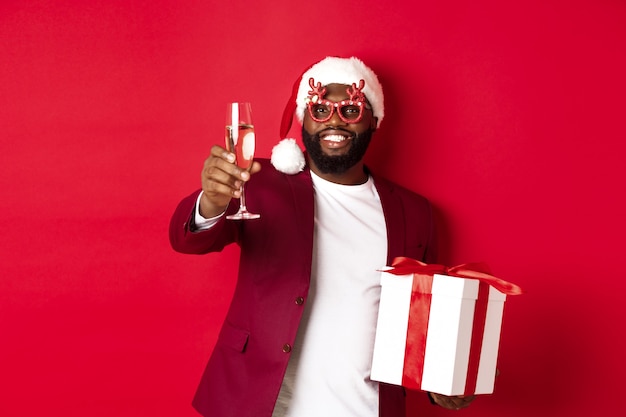 Christmas. Handsome african american man in party glasses and santa hat, holding new year gift and glass of champagne, wishing happy holidays, red background.