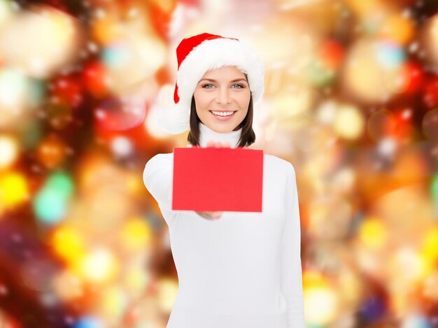 christmas, holdays, people, advertisement and sale concept - happy woman in santa helper hat with blank red card over red lights background