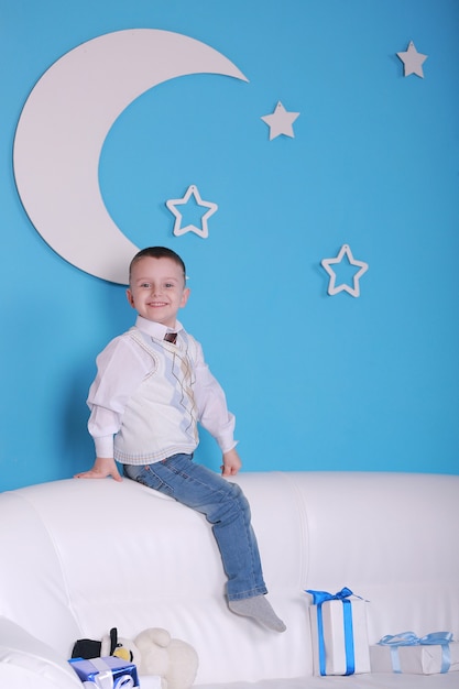 Christmas holiday. little boy on a white sofa with Christmas presents box. blue wall with a white moon on a wall.