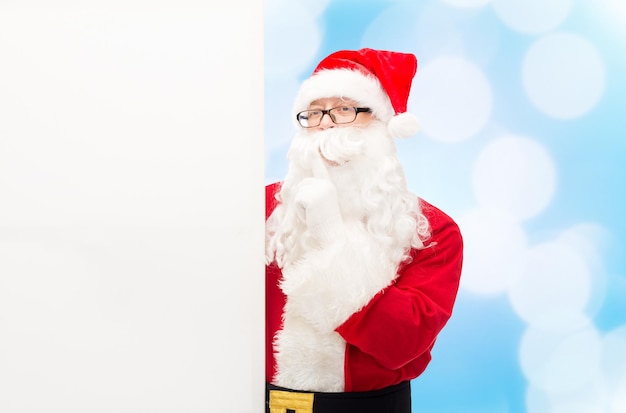 christmas, holidays, advertisement and people concept - man in costume of santa claus with white blank billboard making hush gesture over blue lights background