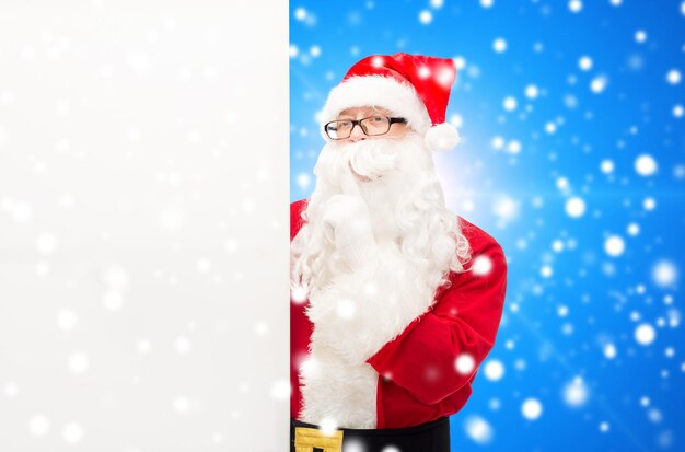 christmas, holidays, advertisement and people concept - man in costume of santa claus with white blank billboard making hush gesture over blue snowy background