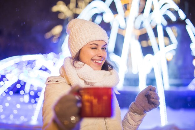 クリスマス休暇。冬の屋外でホットコーヒーのカップと暖かい服を着た美しい笑顔の女性。街の夜。