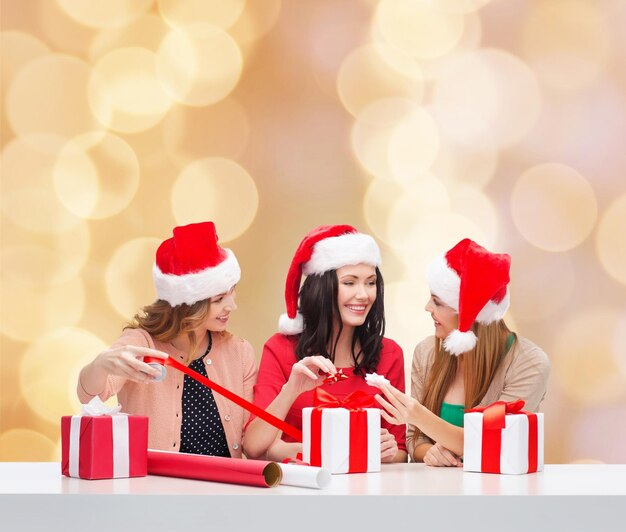 christmas, holidays, celebration, decoration and people concept - smiling women in santa helper hats with decorating paper and gift boxes over beige lights background