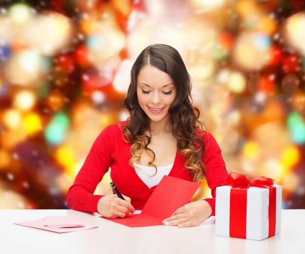 Foto natale, vacanze, celebrazione, saluto e concetto di persone - donna sorridente con scatola regalo che scrive una lettera o invia una cartolina su sfondo a luci rosse