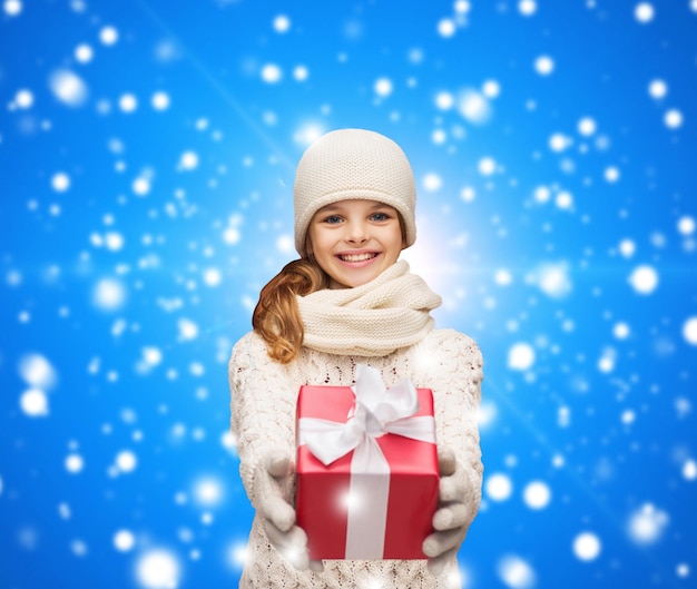 christmas, holidays, childhood, presents and people concept - dreaming girl in winter clothes with gift box over blue snowing background