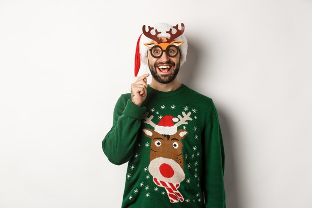 Concetto di natale e festività. felice uomo barbuto che celebra il natale, tiene in mano una maschera da festa e indossa il cappello di babbo natale, sorride stupito, in piedi su sfondo bianco