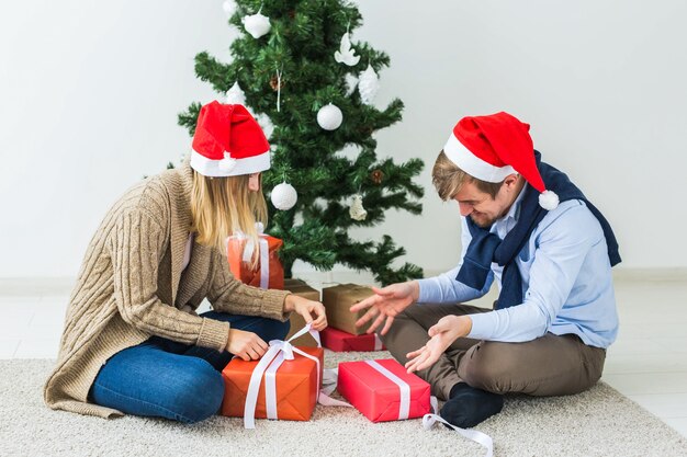 クリスマスと休日のコンセプト-家で贈り物を開くサンタの帽子をかぶっている若い幸せなカップル。