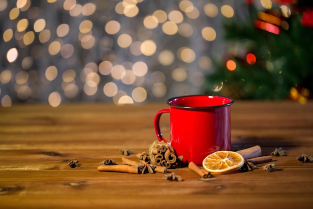 christmas, holidays, cooking and spice concept - close up of tea cup with cinnamon, anise and dried orange on wooden table over lights