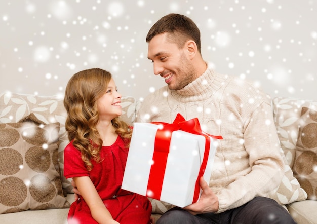 Фото Концепция рождества, праздников, семьи и людей - улыбающиеся отец и дочь держат подарочную коробку дома