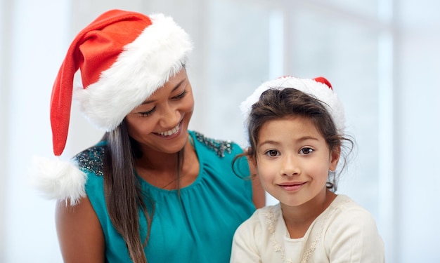 Concetto di natale, vacanze, famiglia, infanzia e persone - madre felice e bambina in cappelli di babbo natale a casa