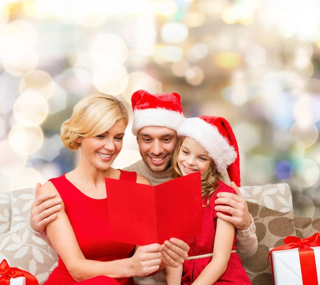 christmas, holidays, family and people concept - happy mother, father and little girl in santa helper hats with gift boxes reading geeting card over lights background