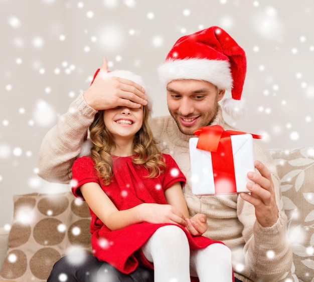 christmas, holidays, family and people concept - smiling father and daughter in santa hats holding gift box and covering eyes at home