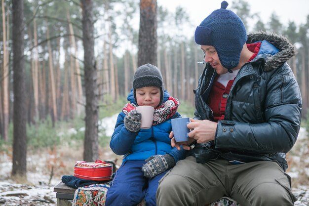 クリスマス休暇、父と息子は熱い新年の飲み物を飲みます。日当たりの良い冬の森の屋外散歩で幸せな家族