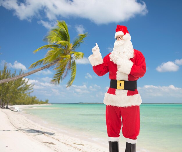 christmas, holidays, gesture and people concept - man in costume of santa claus pointing fingers over tropical beach background