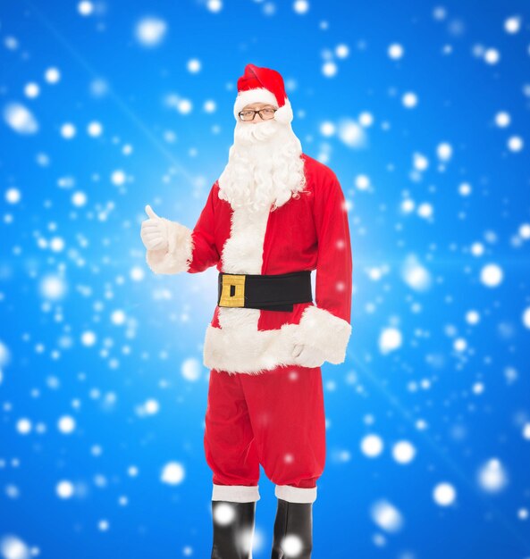 christmas, holidays, gesture and people concept- man in costume of santa claus showing thumbs up over blue snowy background