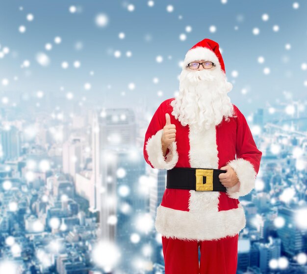 christmas, holidays, gesture and people concept- man in costume of santa claus showing thumbs up over snowy city background