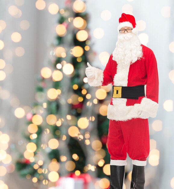 christmas, holidays, gesture and people concept- man in costume of santa claus showing thumbs up over tree lights background