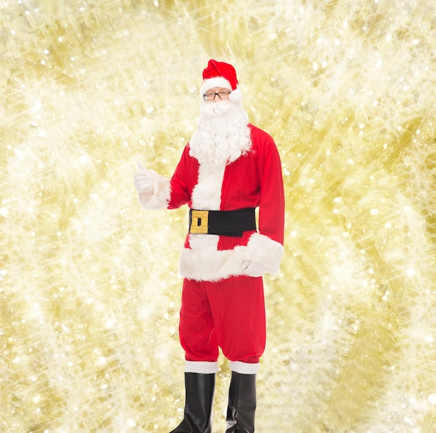 christmas, holidays, gesture and people concept- man in costume of santa claus showing thumbs up over yellow lights background