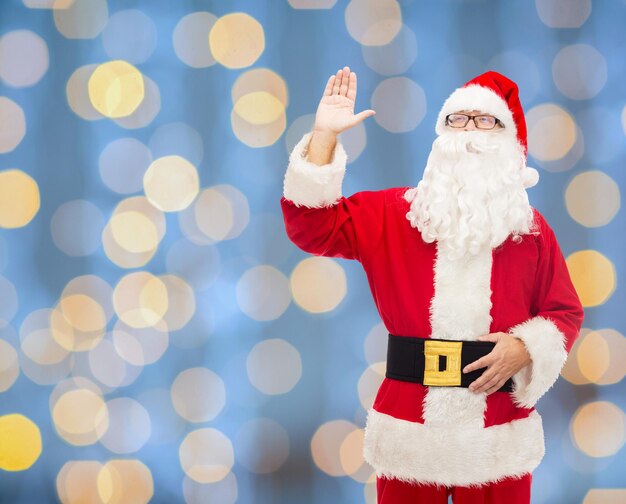クリスマス、休日、ジェスチャー、人々の概念-青い光の背景に手を振っているサンタクロースの衣装を着た男
