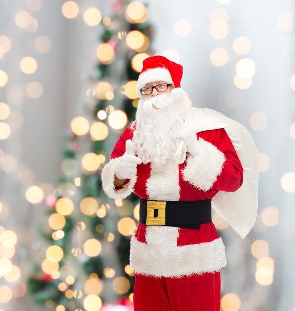 christmas, holidays, gesture and people concept - man in costume of santa claus with bag showing thumbs up over tree lights background