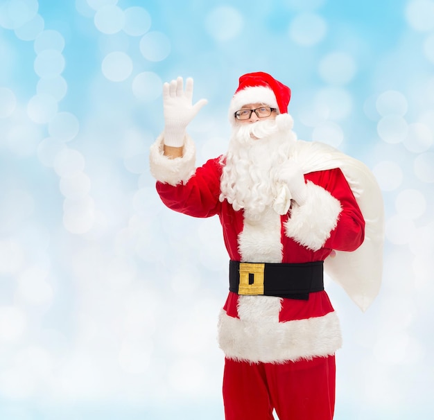 christmas, holidays, gesture and people concept - man in costume of santa claus with bag waving hand over blue lights background