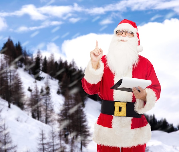 クリスマス、休日、ジェスチャー、人々の概念-雪山の背景の上に人差し指を指しているメモ帳とサンタクロースの衣装を着た男