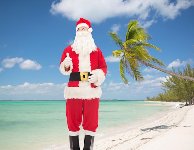 크리스마스, 휴일, 몸짓, 여행, 그리고 사람들 개념 - 열대 해변 배경 위에 엄지손가락을 보여주는 산타클로스 의상을 입은 남자