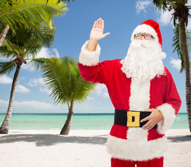 Foto concetto di natale, vacanze, gesto, viaggi e persone - uomo in costume di babbo natale che agita la mano sullo sfondo della spiaggia tropicale