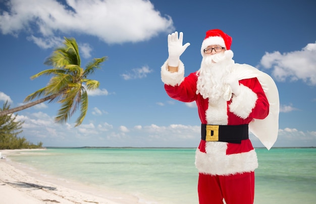 christmas, holidays, gesture, travel and people concept - man in costume of santa claus with bag waving hand over tropical beach background