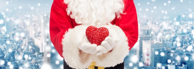 Фото Рождество, праздники, любовь, благотворительность и концепция людей - крупный план санта-клауса с украшением в форме сердца на фоне заснеженного города