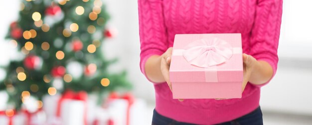 christmas, holidays and people concept - close up of woman in pink sweater holding gift box over living room and tree background