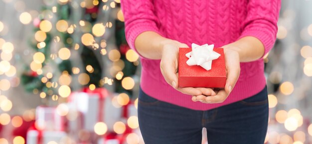 christmas, holidays and people concept - close up of woman in pink sweater holding gift box over tree lights background