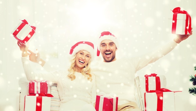 christmas, holidays and people concept - happy couple in santa hats with gift boxes sitting on sofa at home