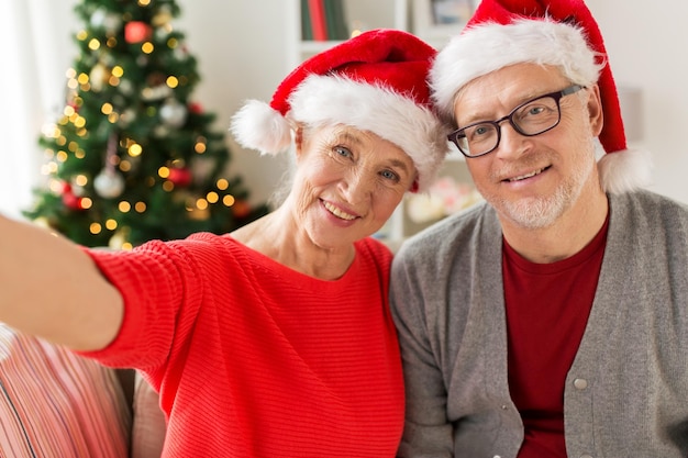 christmas, holidays and people concept - happy smiling senior couple in santa hats taking selfie at home