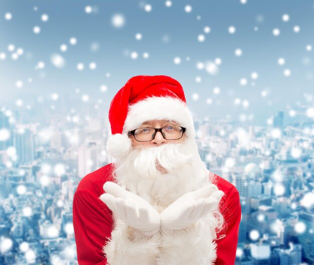 christmas, holidays and people concept - man in costume of santa claus blowing on palms over snowy city background