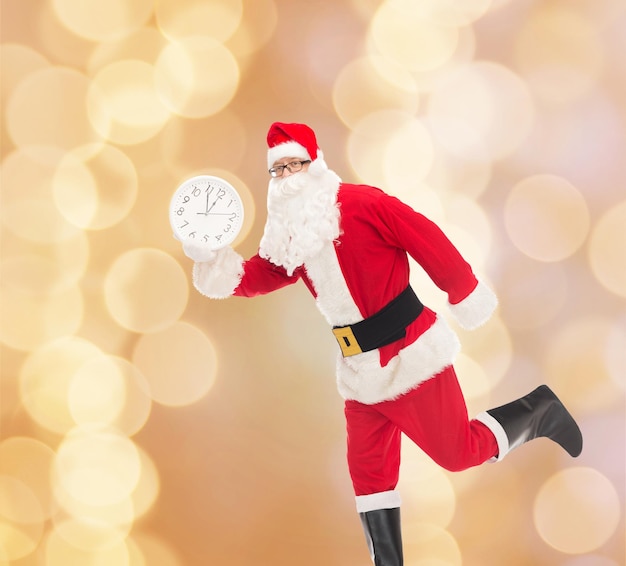 Foto concetto di natale, vacanze e persone - uomo in costume di babbo natale che corre con l'orologio che mostra dodici su sfondo di luci beige