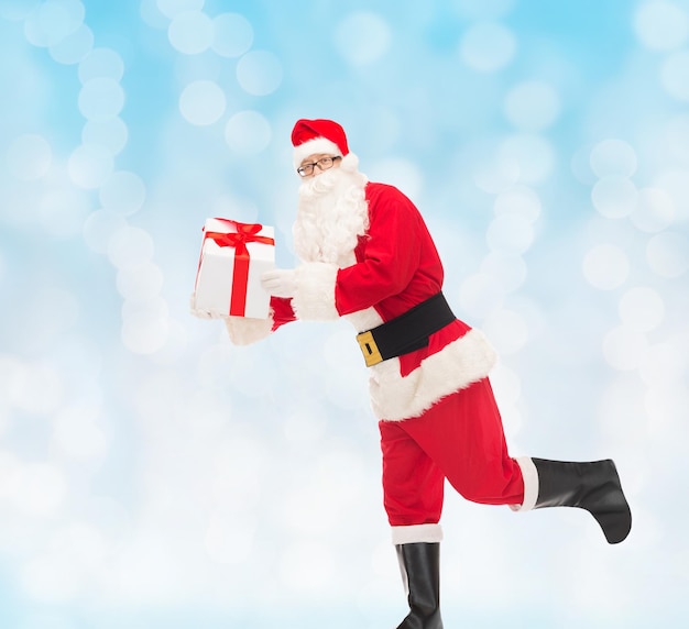 christmas, holidays and people concept - man in costume of santa claus running with gift box over blue lights background