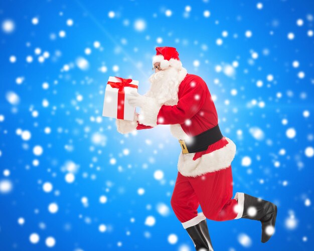 christmas, holidays and people concept - man in costume of santa claus running with gift box over blue snowy background