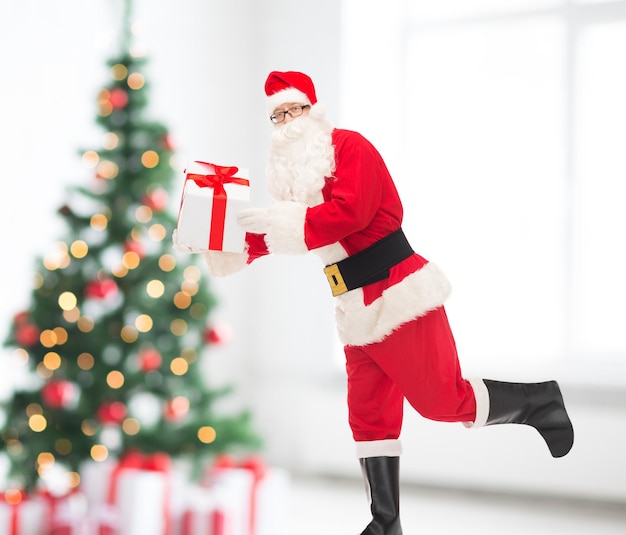концепция рождества, праздников и людей - мужчина в костюме санта-клауса бежит с подарочной коробкой по гостиной и фону дерева