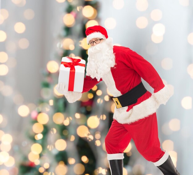 christmas, holidays and people concept - man in costume of santa claus running with gift box over tree lights background