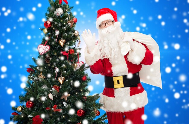 크리스마스, 휴일, 사람 개념 - 산타클로스 복장을 한 남자, 가방을 들고 파란색 눈 덮인 배경 위에 손을 흔드는 크리스마스 트리