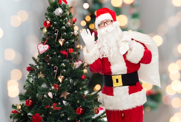 christmas, holidays and people concept - man in costume of santa claus with bag and christmas tree waving hand over lights background