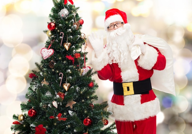 Concetto di natale, vacanze e persone - uomo in costume di babbo natale con borsa e albero di natale che agita la mano sullo sfondo delle luci