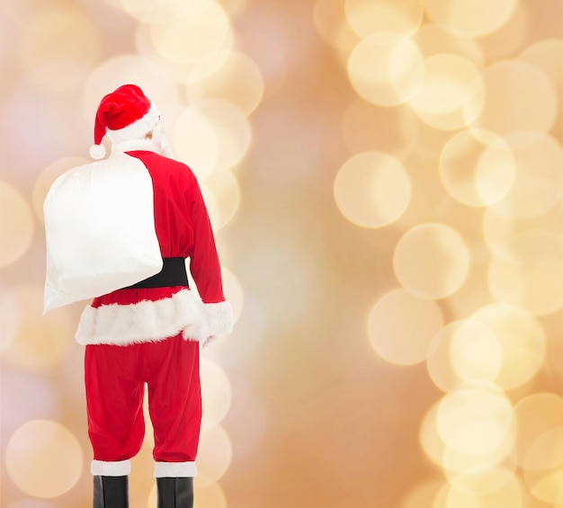 Photo christmas, holidays and people concept - man in costume of santa claus with bag from back over beige lights background