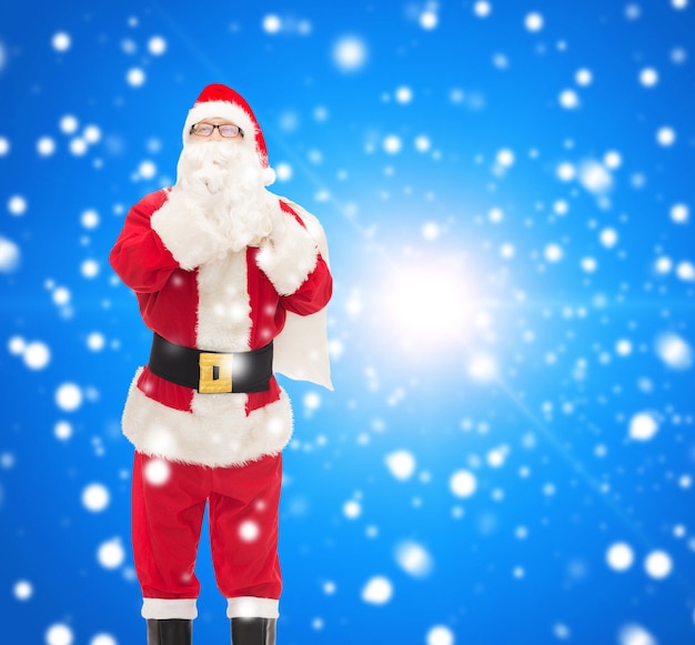 christmas, holidays and people concept - man in costume of santa claus with bag making hush gesture over blue snowy background