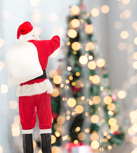 christmas, holidays and people concept - man in costume of santa claus with bag pointing finger from back over yellow lights background over tree lights background