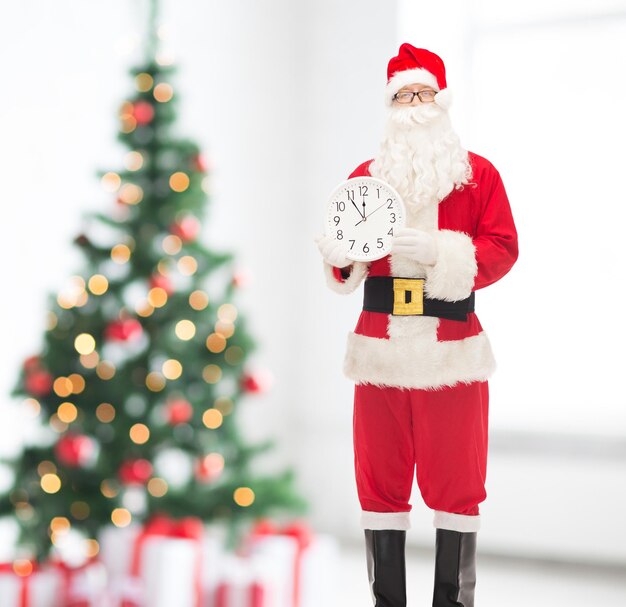 christmas, holidays and people concept - man in costume of santa claus with clock showing twelve over living room and tree background