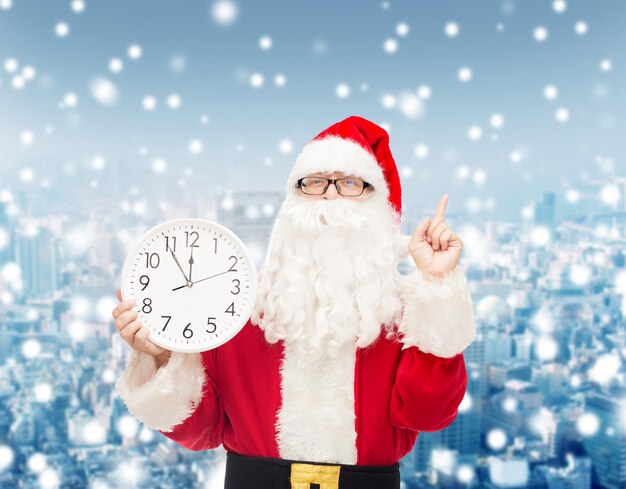 christmas, holidays and people concept - man in costume of santa claus with clock showing twelve pointing finger up over snowy city background
